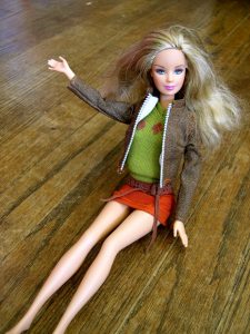 Barbie doll waving, sitting on a hardwood floor, wearing an orange skirt, green top and brown jacket