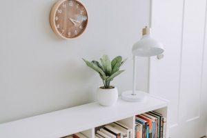 white bookshelf with lamp and plant on it, and a beige clock hanging above it