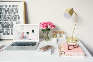 white desk with open laptop, gold lamp, jar of pink roses, pink notebooks with eyeglasses on top