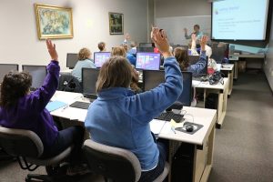 small classroom with adult students behind computers raising their hands