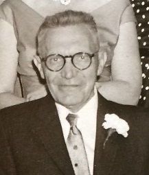 black and white close up of a man wearing glasses, and a suit with a white carnation pinned on it.