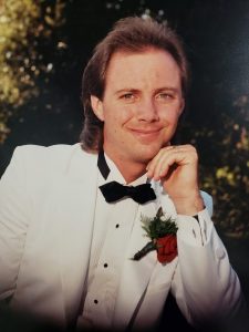 picture of a man in a white tuxedo with a black bow tie and red flower on the lapel
